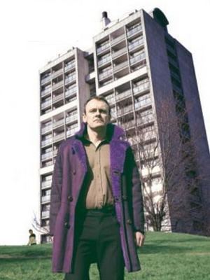 Sean Lock looking afar while wearing a blue coat, brown long sleeves, and black pants in a scene from the 2002 sitcom 15 Storeys High