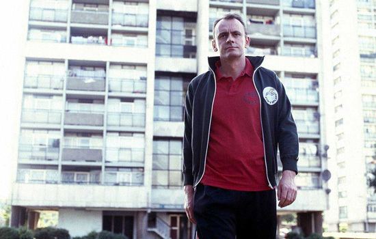 Sean Lock walking while wearing a blue jacket, red t-shirt, and black pants in a scene from the 2002 sitcom 15 Storeys High