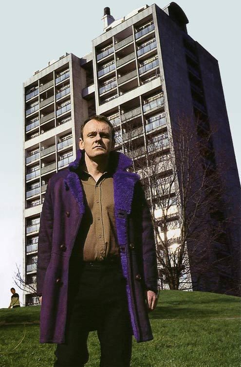 Sean Lock looking afar while wearing a blue coat, brown long sleeves, and black pants in a scene from the 2002 sitcom 15 Storeys High