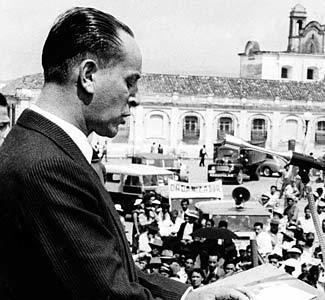 President Jacobo Arbenz Guzman is talking in front of a microphone addressing a crowd of his supporters in 1954. President Jacob is wearing a black coat over white long sleeves.