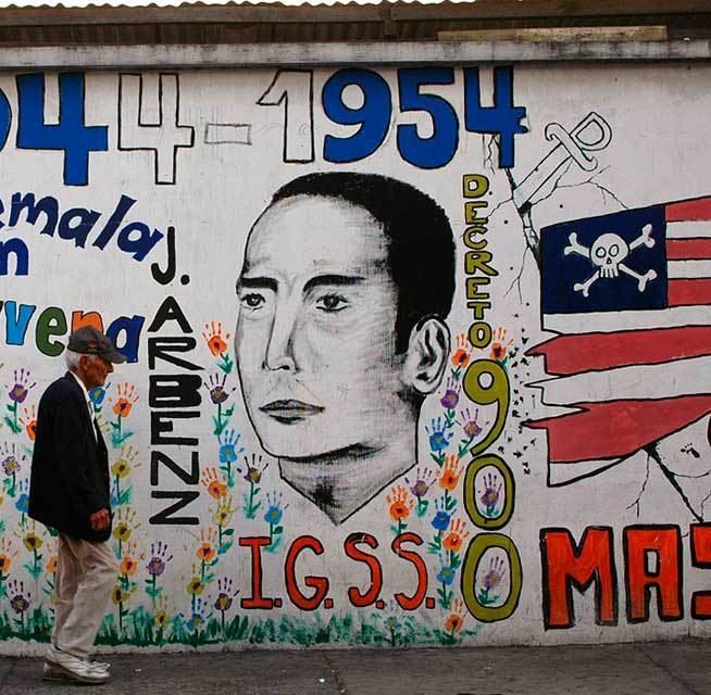 A man walks by a mural of Jacobo Arbenz, Guatemala’s democratically elected president from 1951 to 1954. The man is wearing a black hat, a black coat over white long sleeves, khaki pants, and white shoes.