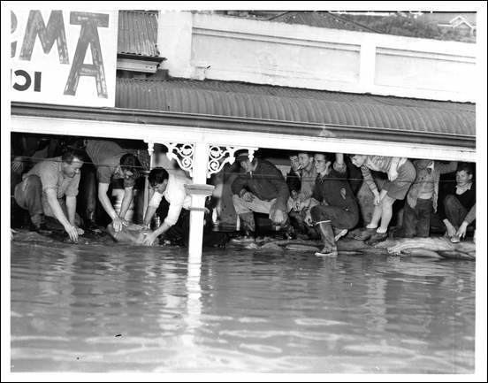 1956 Murray River flood, a group of people under a roof in a flooded street, from left a man is crouching with his left hand down to the water, has black hair wearing gray shirt and black pants, 2nd from left, a man leaning down with his hand holdinga bag from water, has black hair wearing a gray shirt and black pants, 3rd from left, a man crouching, holding a bag from the water, has black hair wearing a white long sleeve, and black pants, 4th from left a man is crouching, has black hair wearing a black long sleeve and gray pants with black shoes, 5th from left a man is smiling crouching, has black hair wearing black shirt, 6th from left a man is smiling, with a cigarette on his mouth has black hair wearing a gray shirt and black pants, 7th from left a man is smiling, crouching, has black hair wearing a black shirt, black pants and black boots, 8th from left, a kid is serious, standing leaning to his right knee with his right hand, has black hair, wearing a striped shirt, black shorts white socks and black shoes, 9th from left a man kneeling holding on the roof with both hands, wearing a white shirt and jacket, black pants, at the right is a man sitting with his knee up in front, has black hair wearing a black long sleeve and black pants with black shoes.