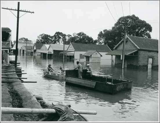 1956 Murray River flood, in black and white has flooded street with electric post from left and right, a flood wall with tubes on top, in the middle is two boat with two person on each boat, from left, a man sitting on the boat rowing with both hands, has black hair, 2nd from left, a man sitting on the boat wearing a long sleeve and gray hat, 3rd a man standing, on a boat with his hand leaning in front panel wearing a black hat and long coat, 4th from left a man is standing, wearing a white hat and long coat, at the right is houses.