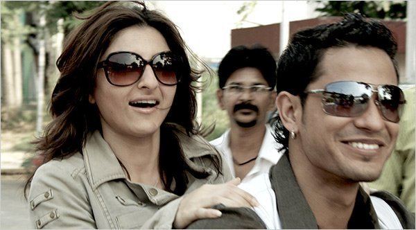 Soha Ali Khan and Kunal Khemu are smiling while riding a motorcycle with a man in the background looking at them in a movie scene from 99, a 2009 Indian Hindi-language crime-comedy film.