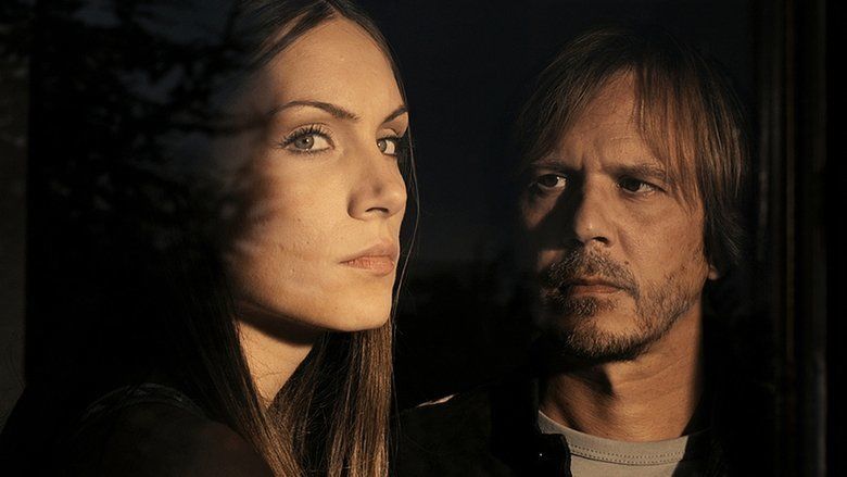 In the movie scene of A Serbian Film 2010, on the left Jelena Gavrilović is serious, looking out the window has long brown hair and brown eyes wearing black shirt, on the right, Srđan Todorović is serious looking at Jelena Gavrilović, has long brown hair, beard and a mustache wearing a black jacket over gray shirt.