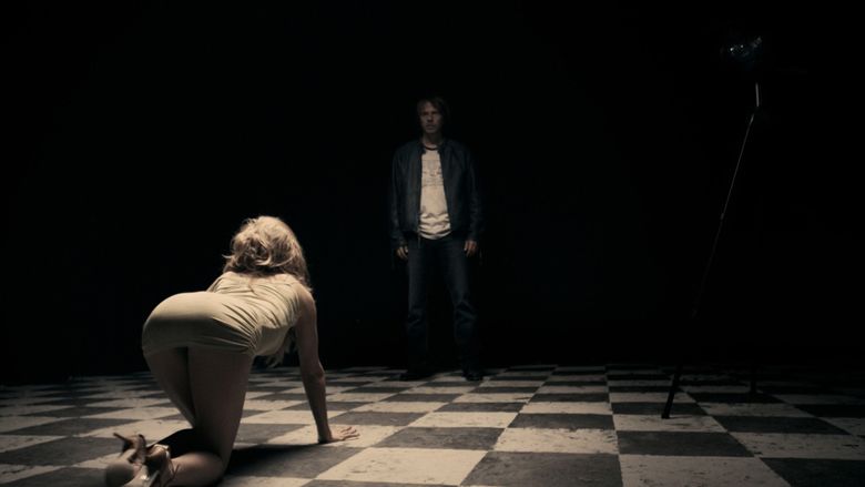 In the movie scene of A Serbian Film 2010, on the left is a woman kneeling with her both hands on the floor while looking at Srđan Todorović, wearing a yellow brown dress and pair of high heels, Srđan Todorović is serious, standing in the middle of a room with a checkered floor and camera on the right with his hands on his side has brown hair beard and a mustache wearing white shirt, black jacket and denim pants with a pair of black shoes.