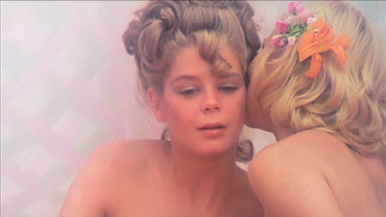 A blonde woman whispering to Kristine De Bell in a scene from the 1976 American musical fantasy adult film, Alice in Wonderland
