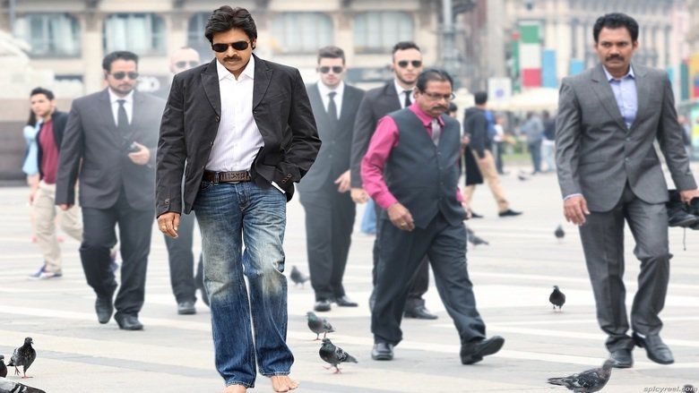 Pawan Kalyan as Goutham Nanda looking on the ground while his hand on his pocket and on his back are men wearing suits. Pawan is with a mustache, wearing sunglasses, a black coat over a white shirt, and blue pants in a movie scene from Attarintiki Daredi, a 2013 Indian Telugu-language action drama film.