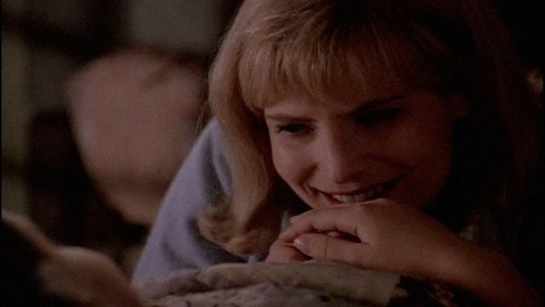 Jennifer Jason Leigh as Anney smiling and holding Jena's hand (Ruth) in a scene from a 1996 American drama film, Bastard Out of Carolina. Jennifer has short blonde hair with bangs wearing a purple top.