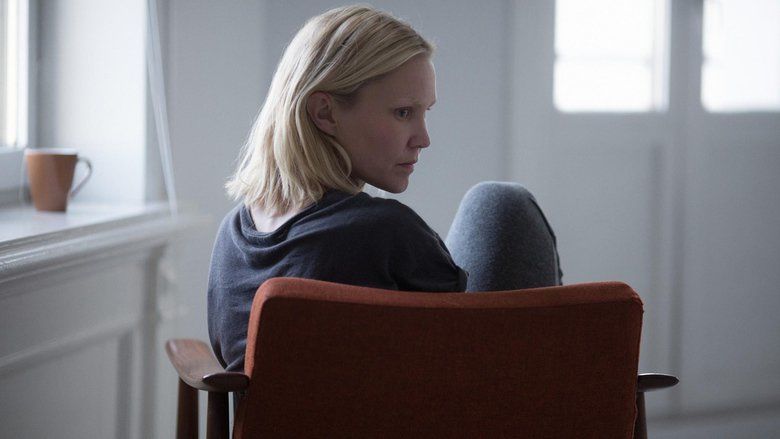 In the movie scene of Blind(2014), In a white room with a large glass wooden door, Ellen Dorrit Petersen is serious, sitting on a maroon wooden chair, with a brown cup on the window on the left, has blonde hair, wearing a black shirt and gray pants.