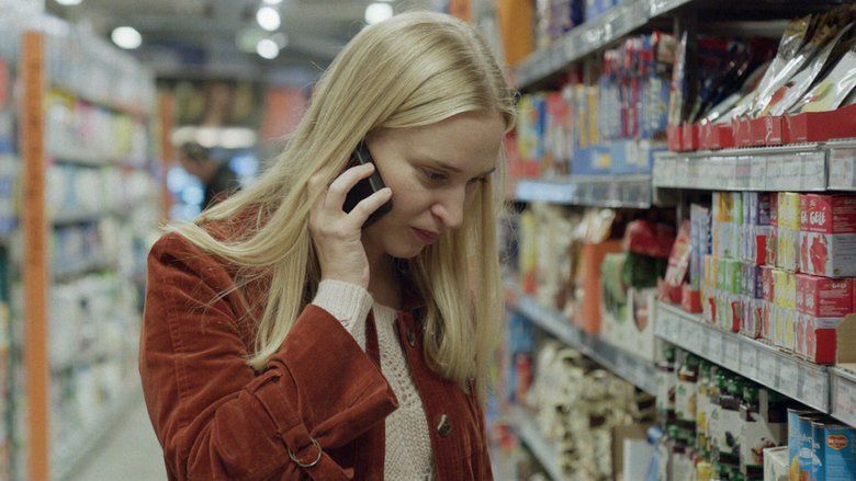 In the movie scene of Blind(2014), In a supermarket aisle, Vera Vitali is serious, looking down, standing while holding the phone in her ears with her right hand, has long blond hair wearing a white crochet long sleeve under a brown coat.
