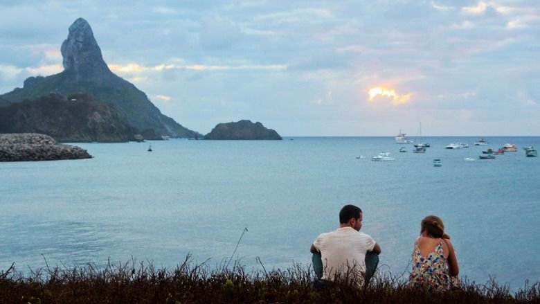 A man and a woman looking afar while sitting on the grass near the sea where they can watch the sunset, hills, and boats in a scene from the 2014 Brazilian drama film, Blue Blood
