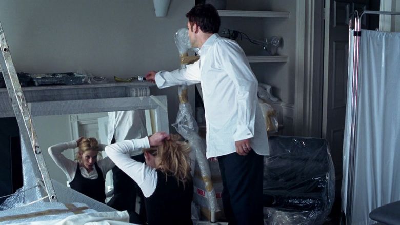 In the movie scene of Closer 2004, in a room with white walls, shelves and things wrapped with plastic and cloth, from left Julia Roberts is serious, sitting in front of a mirror, tying her hair, has blond hair wearing white long sleeve under black tank top at the right, Clive Owen is standing, back view, reaching towards the shelves, has black hair wearing a white long sleeves a ring and black pants.