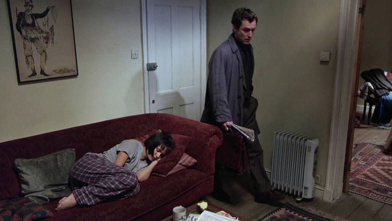 In the movie scene of Closer 2004, in a room, has white walls with hanging painting, a brown sofa with four throw pillows, from left a red checkered, gray, maroon and a red striped throw pillow, a brown table with a cup and papers on top, and a white heater, from left, Natalie Portman is sleeping, laying on the sofa, has brown short hair wearing a gray shirt and checkered pajama, at the right Clive Owen is serious, looking down, holding a news paper, has black hair wearing a black coat over a black shirt with a black pants and shoes.