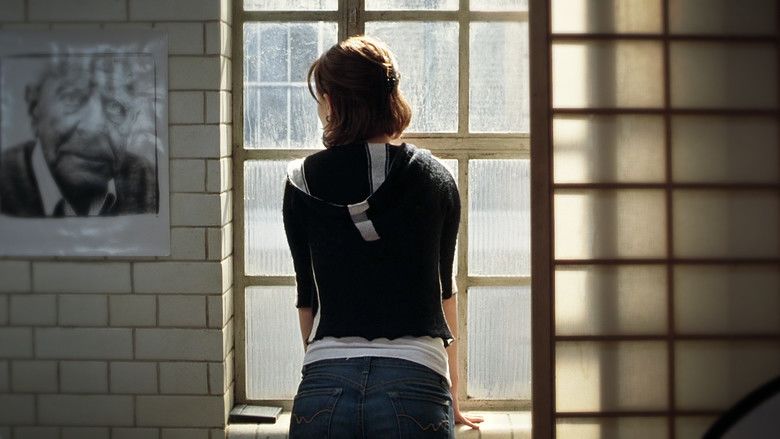 In the movie scene of Closer 2004, in a room, has white brick walls with a photo hanging on the wall and a white window,Natalie Portman is standing, back view, has black short hair wearing a white tank top under a black jacket and denim pants.