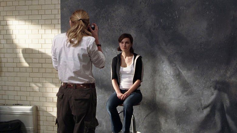 In the movie scene of Closer 2004, in a room, has white brick walls, and a gray backdrop,From left, Julia Roberts is standing, back view, taking a photo of Natalie, has blond hair wearing a white long sleeve with black bracelet and a black pants, Natalie Portman(right) is serious, sitting on a bar stool, has black short hair wearing a white tank top under a black jacket and a denim pants.
