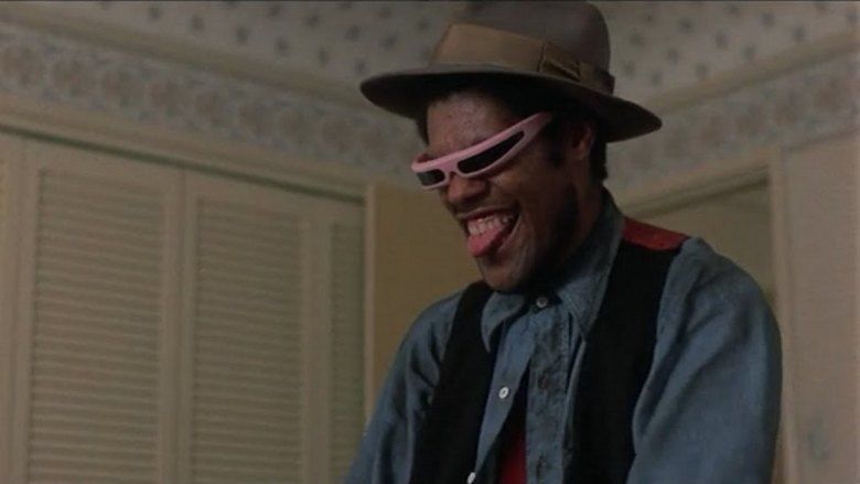 Laurence Fishburne as Cutter looking happy with his tongue out in a scene from the 1982 film Death Wish II, wearing a red shirt under a blue long-sleeved shirt and a black vest, pink sunglasses, and a brown hat