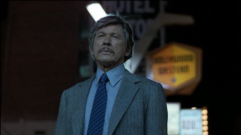 Charles Bronson as Paul Kersey looking serious with signages in the background, having a gray hair and a mustache, wearing a blue shirt, a necktie with blue and white stripes, and a gray coat