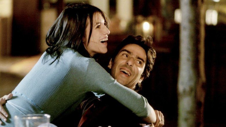 Charlotte Gainsbourg (left) smiling, has long black hair, both hands hugging Yvan Attal (right) wearing a blue sweater with a cup of glass on the table. Yvan Attal is smiling and has black hair, holding Charlotte’s waist with his right hand, wearing a black jacket.