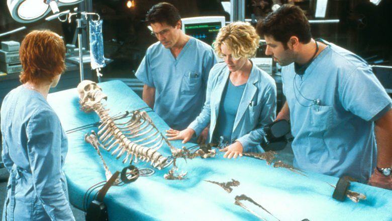 Margot Rose, Kevin Bacon, Elisabeth Shue, and Greg Grunberg (from left to right) are looking at the human skeleton in an operating room and all of them are wearing blue scrub suits in a movie scene from Hollow Man, a 2000 science fiction thriller film.