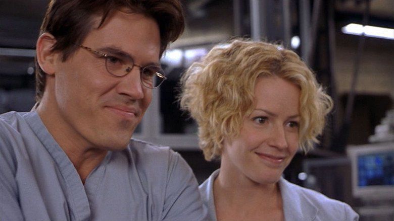 Josh Brolin and Elisabeth Shue with tight-lipped smiles while looking at something. Josh is wearing eyeglasses while Elisabeth with blonde hair and both are wearing gray scrub suits in a movie scene from Hollow Man, a 2000 science fiction thriller film.
