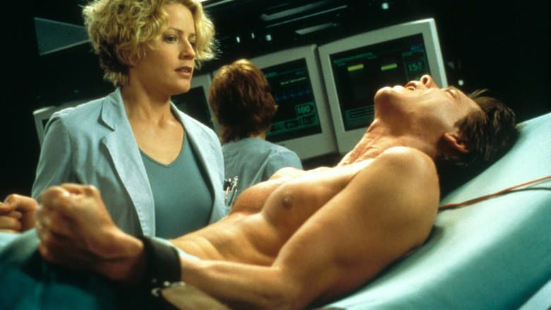 Elisabeth Shue with a worried face while looking at a man lying in an operating bed with Margot Rose at the back looking at the monitors in a movie scene from Hollow Man, a 2000 science fiction thriller film.