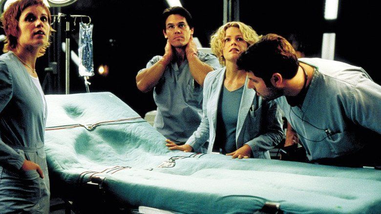 Margot Rose, Kevin Bacon, Elisabeth Shue, and Greg Grunberg (from left to right) with serious faces while looking at something in an operating room and all of them are wearing blue scrub suits in a movie scene from Hollow Man, a 2000 science fiction thriller film.