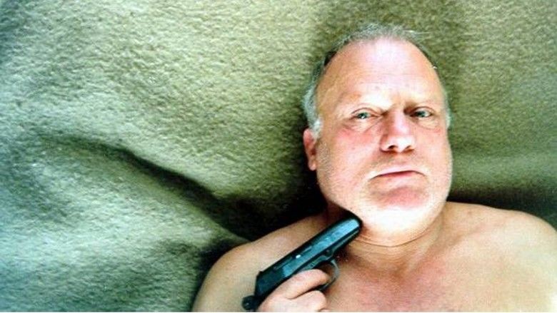 Philippe Nahon seriously looking and laying on a cream color blanket while holding a gun pointing at his neck, he had gray hair and topless