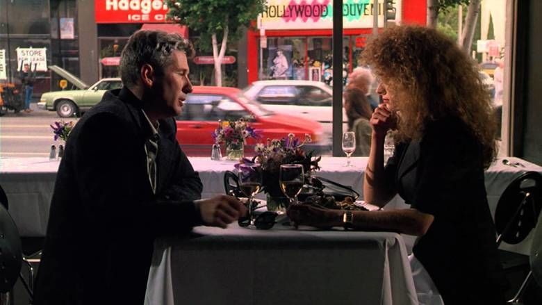 Richard Gere and Nancy Travis talking and looking at each other while holding a glass of wine in the restaurant, with cars passing by in their background, in a scene from the 1990 American crime thriller film, Internal Affairs. Richard is wearing a black coat and Nancy is wearing a wristwatch and a black dress.