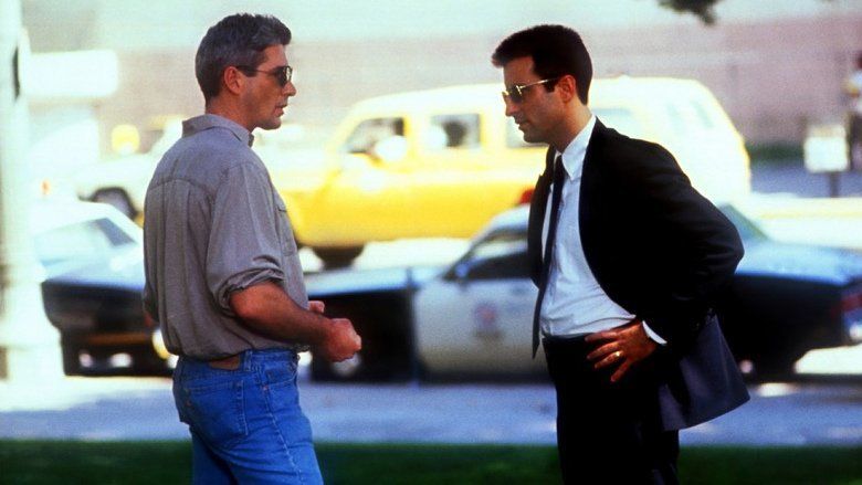 Richard Gere and Andy Garcia talking to each other with serious faces and there are cars in the background in a scene from the 1990 American crime thriller film, Internal Affairs. Richard is wearing a sunglasses, a gray polo shirt, and denim pants while Andy is wearing sunglasses, a ring, black pants, and a white long sleeve under a black necktie and a black coat