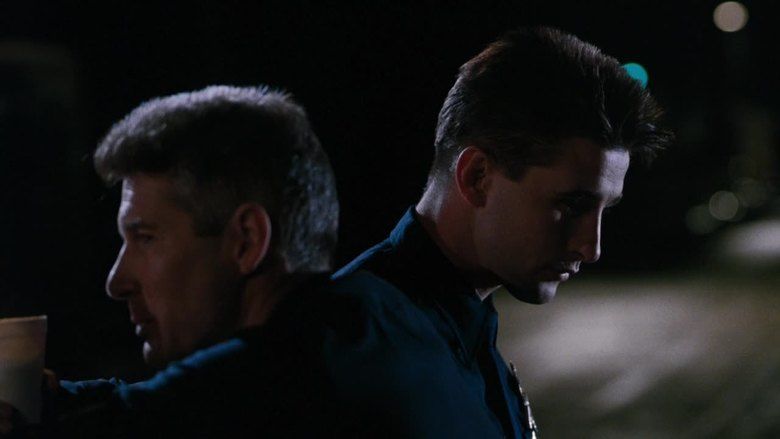 Richard Gere and William Baldwin are looking afar in the dark with serious faces in a scene from the 1990 American crime thriller film, Internal Affairs. Richard (left) is holding a white styrofoam cup and both of them are wearing blue long sleeves