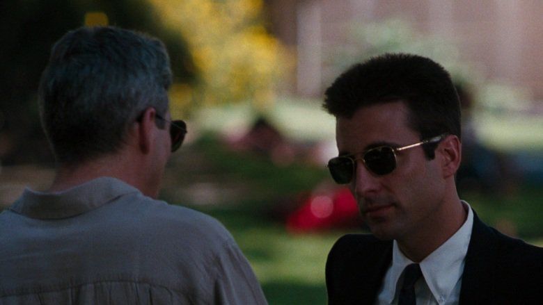 Andy Garcia looking at the man in front of him with a serious face in a scene from the 1990 American crime thriller film, Internal Affairs. Andy is wearing sunglasses and a white long sleeve under a black necktie and black coat while the man in front of him is wearing a gray polo shirt and sunglasses