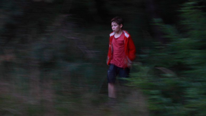 The movie scene of Jack (2014 film),  in the middle of a forest with green leaves and bushes, Ivo Pietzcker is serious, standing, mouth half open, with his hand in his pocket, has black hair wearing a red shirt under a red jacket and blue shorts.