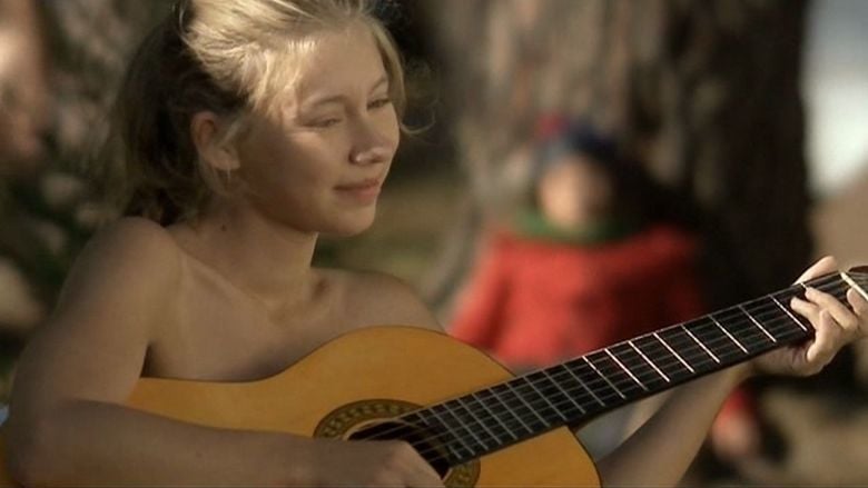 Géraldine Martineau is smiling while playing the guitar with a boy in the background from a movie scene of La Fonte des Neiges, a 2009 short comedy-drama film. Geraldine is topless while the boy is wearing a beanie, a green scarf, and a red jacket.
