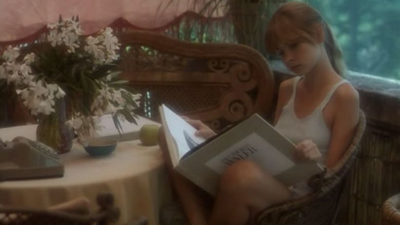 In the Laura (1979 film) movie scenes, Laura is serious, has brown hair, sits down and reads a book on a brown chair on the terrace, with a brown big chair on her right, a pink curtain, and a pink table in front with a flower vase and white flower, a telephone, a blue bowl, and a green apple. She is wearing a white sleeveless top.