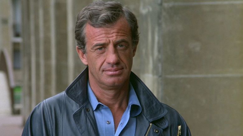 Jean-Paul Belmondo as Josselin Beaumont dit 'Joss' with a serious face and wearing a black jacket over a blue polo shirt in a movie scene from Le Professionnel, a 1981 French action thriller film.