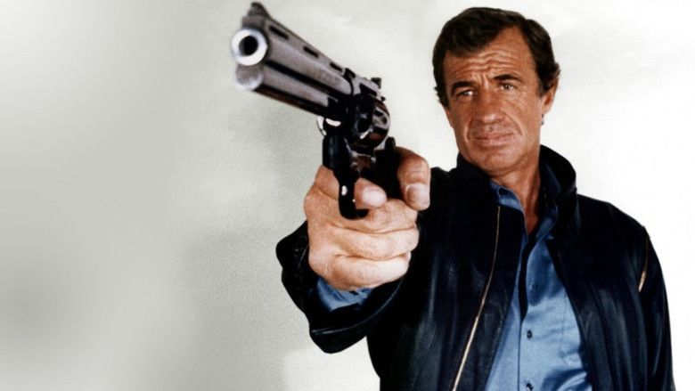 Jean-Paul Belmondo as Josselin Beaumont dit 'Joss' with a serious face while holding a gun and wearing a black jacket over a blue polo shirt in a movie scene from Le Professionnel, a 1981 French action thriller film.
