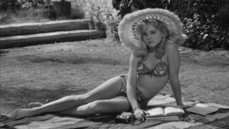 Sue Lyon as Lolita with a serious face while sitting on the ground, wearing a fur hat, and a floral two-piece swimsuit in a movie scene from Lolita, a 1962 psychological comedy-drama film.