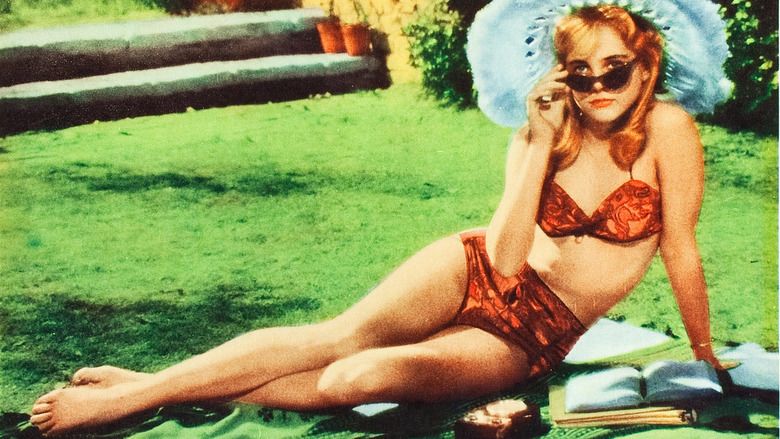 Sue Lyon as Lolita with a serious face while sitting on the ground, wearing sunglasses, a fur hat, and a red two-piece swimsuit in a movie scene from Lolita, a 1962 psychological comedy-drama film.