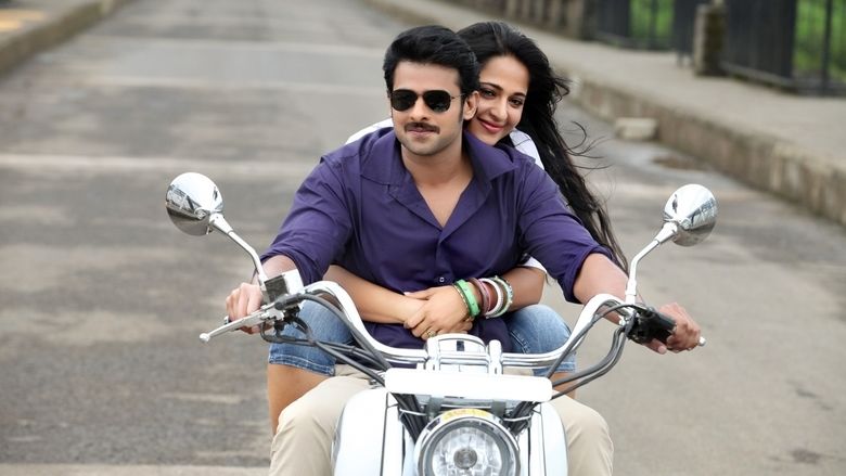Prabhas and Anushka Shetty smiling while riding on the motorcycle in a scene from the 2013 action drama film, Mirchi