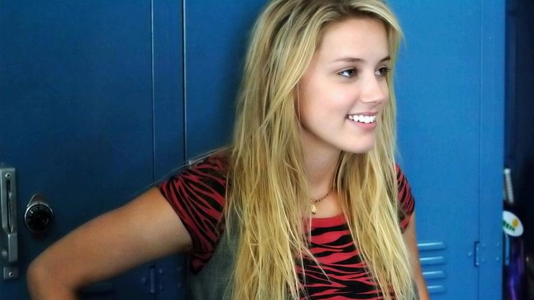 Amber Heard smiling at someone while leaning on a blue student locker, she has long blonde hair, wearing a necklace and a black and red shirt