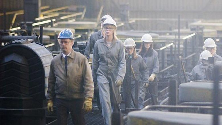 The man in front is serious and wears yellow gloves on both hands, a blue hard hat, white long sleeves with a blue necktie under a brown jacket, and black pants. 2nd from left is Josey Aimes, who has blonde hair, is serious, and wears gray gloves, a white hard hat, a gray protective suit, and eyeglasses. 3rd from left is a man, wearing a gray protective suit, and a white hard hat. 4th from left is a woman, who has blonde hair, is serious, and wears gray gloves, a white hard hat, a gray protective suit, and eyeglasses.5th from left is a woman, serious, has black hair, and wears gray gloves, a white hard hat, a gray protective suit, and eyeglasses. On right is a person wearing eyeglasses, white hard hat and a gray protective suit.