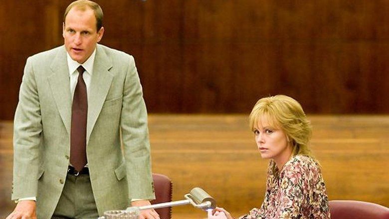 Woody Harrelson (left) is serious, mouth half open, both hands on the table, has blonde hair, wears white long sleeves, a brown necktie under an off-white suit, and pants with a black belt. On right is Charlize Theron, sitting down on a red chair, has blonde hair, both hands holding each other, are placed on the table, and has a roller in front, she is wearing a floral long-sleeve dress.