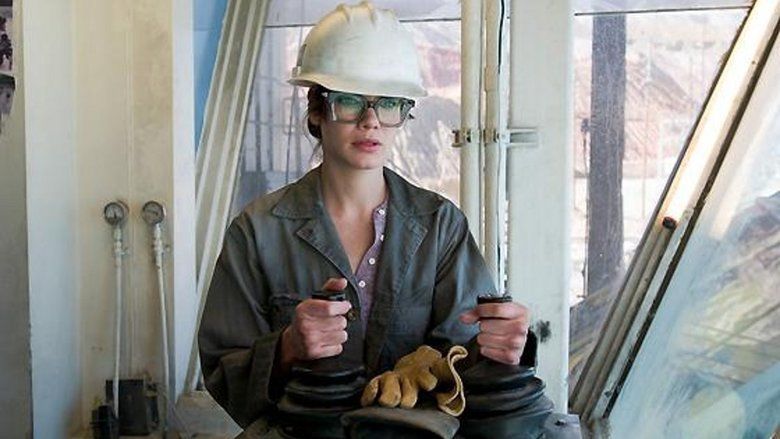 Josey Aimes is serious, both hands holding a steering wheel, has two yellow gloves in front, wears eye protectors, a white hard hat, and a violet shirt under a gray protective suit.