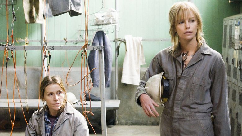 Frances McDormand (left) is serious, has blonde hair, looks to her left, wears a violet shirt, and a blue jacket under a gray suit, and behind her are ropes and clothes. Charlize Theron (right) is serious, has blonde hair with bangs, her right hand holding a white hard hat, has eyeglasses on her chest, wears a gray suit, and on her left is a cabinet.