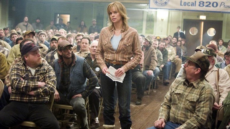 Josey Aimes (middle) is serious, has blond hair, both hands holding a piece of paper, and wears a blue shirt under brown long sleeves, denim pants, and brown boots. On both of her sides are men looking at her, sitting down.