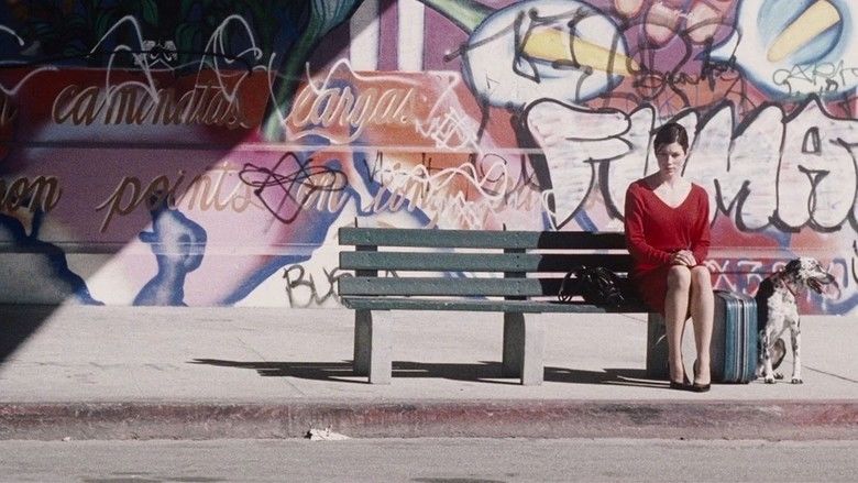 Jessica Biel sitting on a gray bench and beside her is a blue suitcase and a black and white dog looking afar while his tongue is out, and a wall mural at the back. Jessica is wearing a red long sleeve dress and black heels in a scene from the 2008 drama film, Powder Blue
