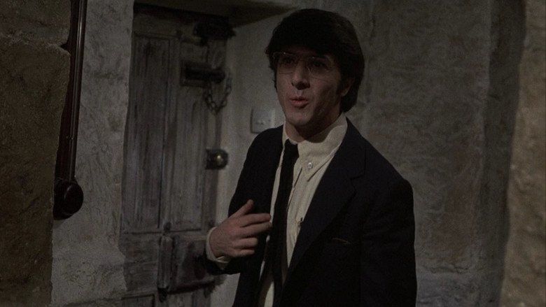 Dustin Hoffman as David Sumner is talking while pointing himself, wearing eyeglasses, black coat over white long sleeves, and black tie in a movie scene from Straw Dogs, a 1971 psychological thriller film.