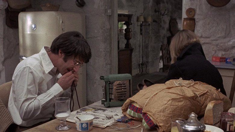 Dustin Hoffman biting a rope, wearing eyeglasses and white long sleeves with a woman showing her back, and wearing a black coat in a movie scene from Straw Dogs, a 1971 psychological thriller film.