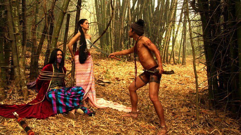 Ananda Everingham, Chermarn Boonyasak, and Dom Heatrakul (from left to right). Ananda was tied by a rope and sitting on the ground with a mouth covering, Chermarn is standing with her hands tied on a bamboo tree, while Dom is standing and holding his sword pointed at Chermarn with bamboo trees in the background from a movie scene of The Outrage, a 2011 Thai Drama movie. Ananda is wearing an army green shirt over a long maroon cardigan and a tri-colored skirt. Chermarn is wearing a white tube top and a pink printed skirt while Dom has body tattoos, and a cloth covering his head and is topless wearing printed short tights.